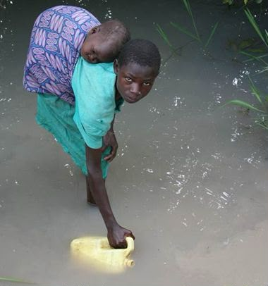 A young girl carrying a baby while filling a water container from the river